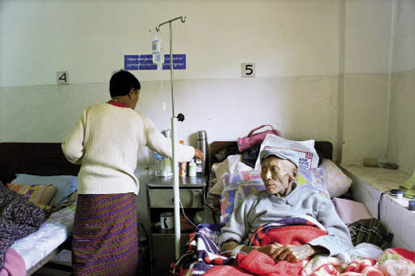 Een oude man met pet op aan het infuus in bed op zaal; zijn vrouw zet spullen op zijn kastje.